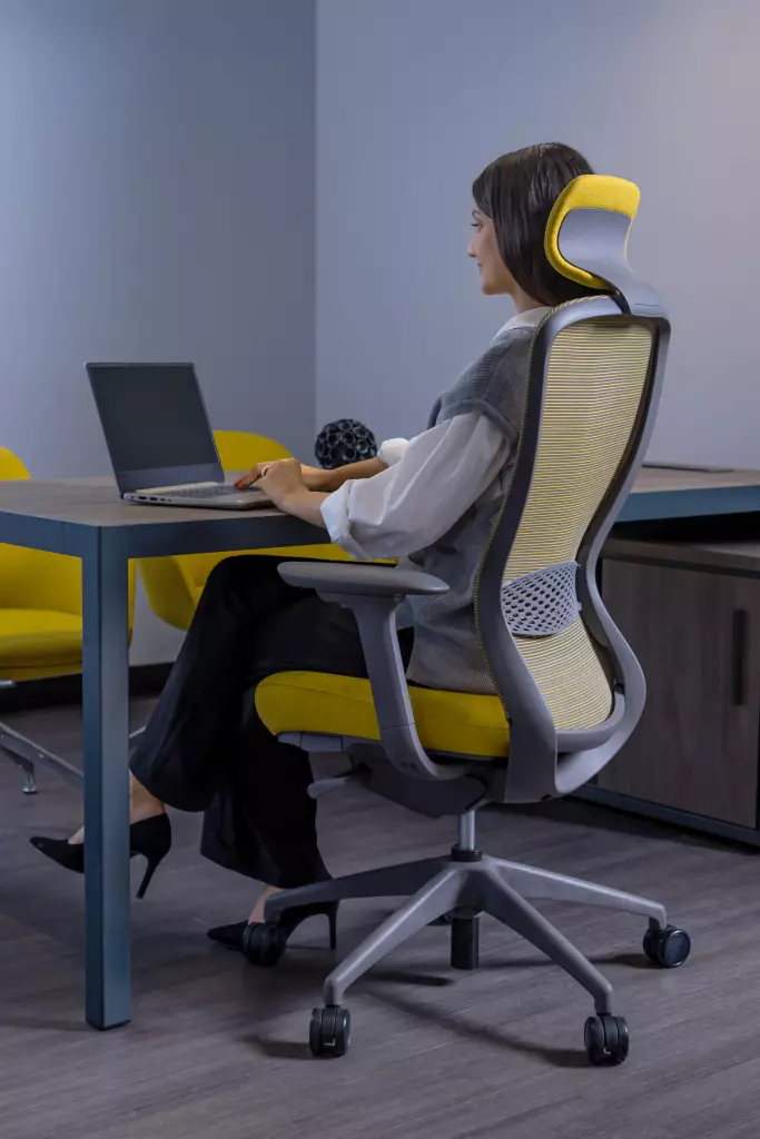 A woman sitting in a modern ergonomic office chair with a yellow and gray design, working on a laptop at a sleek desk.